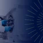 woman with long dark hair wearing a white lab coat and blue rubber gloves removes trays from an autoclave